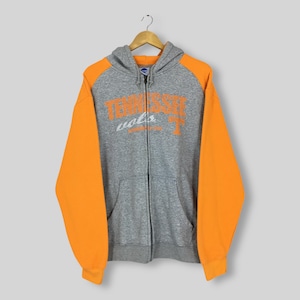 May include: A gray zip-up hoodie featuring bright orange sleeves and a hood. The front displays the text Tennessee Vols in orange and white lettering with a small orange T logo. It includes a full-length front zipper and two front pockets.