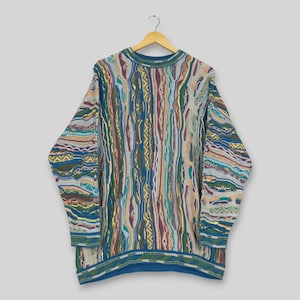 Könnte beinhalten: Ein langärmeliger Pullover mit Rundhalsausschnitt und einem komplexen, mehrfarbigen, strukturierten Strickmuster. Das Design umfasst vertikale Streifen, wellenförmige Linien und geometrische Formen in Blau-, Grün-, Gelb-, Beige- und Burgundertönen.