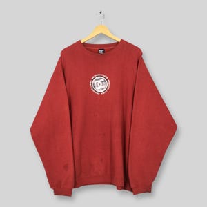May include: A red DC Shoes sweatshirt with a circular logo on the chest. The logo is white and silver with the words "DC SHOECO USA" around the perimeter. The sweatshirt is hanging on a wooden hanger.