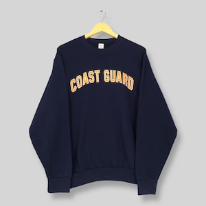 May include: Navy blue sweatshirt with the words "COAST GUARD" in arched orange lettering. The sweatshirt is hanging on a yellow hanger against a light gray background. The sweatshirt has long sleeves and a crew neck.