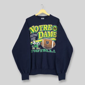 May include: A navy blue long-sleeve sweatshirt featuring a graphic print that reads NOTRE DAME FIGHTING IRISH FOOTBALL in yellow and green lettering. The design includes an illustration of a leprechaun mascot and a brown football with an ND logo.