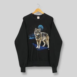 May include: Black sweatshirt with a detailed wolf illustration under a starry night sky and a blue moon. The wolf is in brown and grey, standing on a blue surface. The design includes a forest silhouette. The sweatshirt is hanging on a wooden hanger.