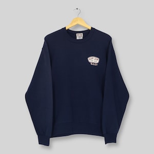 May include: A navy blue sweatshirt featuring the text "RON JON SURF SHOP U.S.A." in a white and red oval design. The sweatshirt is suspended from a wooden hanger.