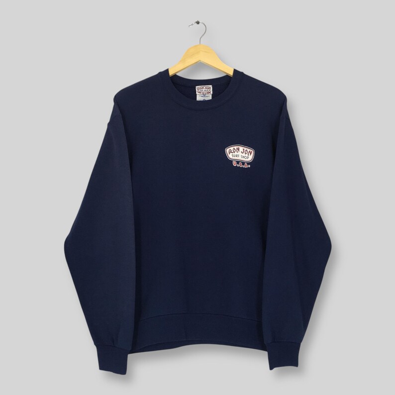 May include: A navy blue sweatshirt featuring the text "RON JON SURF SHOP U.S.A." in a white and red oval design. The sweatshirt is suspended from a wooden hanger.