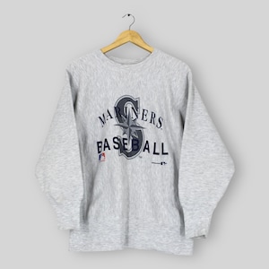 May include: A light gray Mariners baseball sweatshirt with the team's logo in navy blue and white. The logo includes the word "MARINERS" above "BASEBALL" and a compass rose design. The MLB logo is visible on the bottom left.