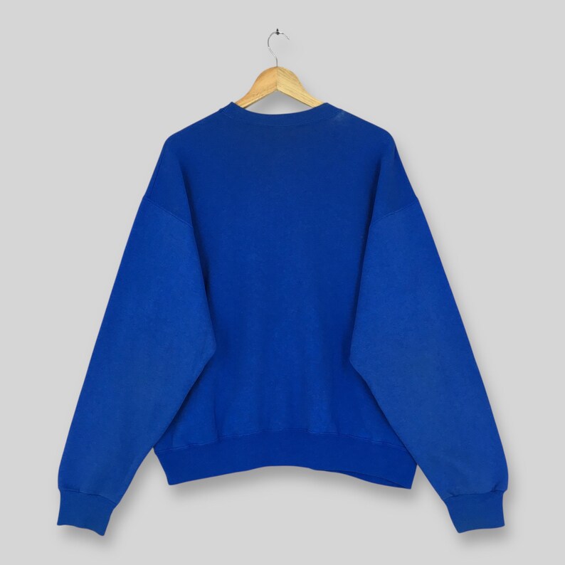 May include: A royal blue sweatshirt with long sleeves, hanging on a wooden hanger. The sweatshirt is a solid color and appears to be made of a soft material. The background is a light gray.