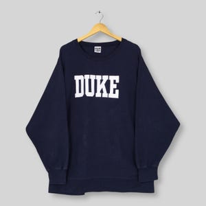Può includere: Felpa blu navy con la scritta "DUKE" a grandi lettere bianche sul petto. La felpa ha un girocollo e maniche lunghe. L'indumento è appeso a una gruccia di legno.