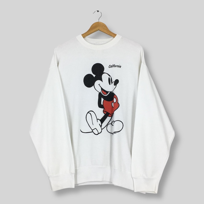 May include: White sweatshirt featuring a large graphic of Mickey Mouse in a classic pose, with red shorts and black ears. The word "California" is printed above the character. The sweatshirt is hanging on a wooden hanger.