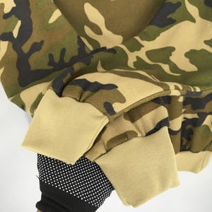 May include: Close-up of a camouflage-patterned sweatshirt. The fabric features a mix of green, brown, and tan hues. The cuffs and bottom hem are a solid tan color. A black and white dotted glove is visible.
