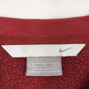 Może przedstawiać: Zbliżenie czerwonego kołnierza bluzy z białą metką. Na metce znajduje się logo Nike i napis "MADE IN SINGAPORE FABRIQUE A SINGAPOUR". Bluza jest w jednolitym czerwonym kolorze.
