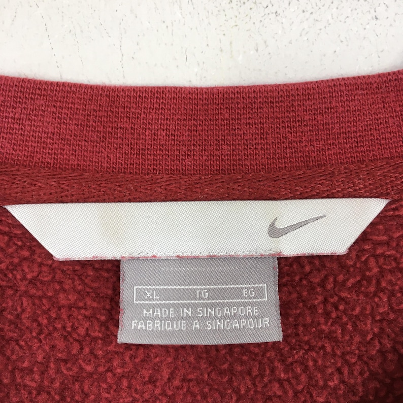 Może przedstawiać: Zbliżenie czerwonego kołnierza bluzy z białą metką. Na metce znajduje się logo Nike i napis "MADE IN SINGAPORE FABRIQUE A SINGAPOUR". Bluza jest w jednolitym czerwonym kolorze.
