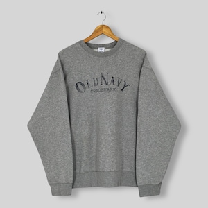 May include: A heather grey crewneck sweatshirt featuring long sleeves and a ribbed collar, cuffs, and hem. The chest displays the text OLD NAVY TRADEMARK in a distressed, dark charcoal font. The garment is displayed on a wooden hanger.