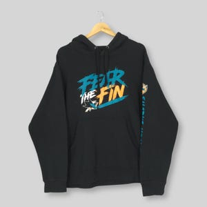 May include: Black hoodie with the text "FEAR THE FIN" in teal and orange. The hoodie features a front pocket and a hood with a drawstring. The sleeve has a teal and orange design. The hoodie is hanging on a wooden hanger.