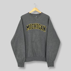 Könnte beinhalten: Ein graues Sweatshirt mit Rundhalsausschnitt und dem Wort "MICHIGAN" in gelben Blockbuchstaben mit marineblauer Umrandung. Das Sweatshirt hängt an einem Holzkleiderbügel.
