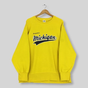 Peut inclure: Un sweat-shirt à col rond et manches longues jaune vif avec l'inscription The University of Michigan en lettres cursives bleu marine foncé sur la poitrine. Le vêtement présente un col, des poignets et un ourlet côtelés, suspendu à un cintre en bois.