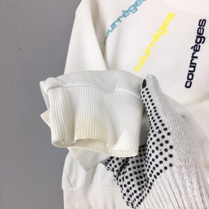 May include: White sweatshirt with the word "courr&egrave;ges" in blue, yellow, and black lettering. The sleeve cuff is ribbed and shows some discoloration. A white glove with black dots is in the foreground.