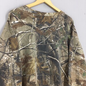Op de afbeelding: Een camouflage shirt met lange mouwen in bruin-, groen- en beigetinten, met een realistisch boom- en takkenpatroon. Het shirt hangt aan een houten hanger tegen een lichtgrijze achtergrond.
