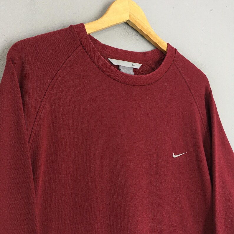 Może przedstawiać: Bordowa bluza z okrągłym dekoltem i małym białym logo Nike na piersi. Bluza wisi na drewnianym wieszaku. Rękawy mają kr&oacute;j raglan.