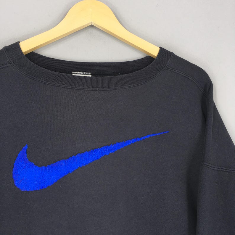 Op de afbeelding: Zwart sweatshirt met ronde hals en een groot blauw Nike swoosh-logo. Het sweatshirt hangt aan een houten hanger tegen een grijze achtergrond. Het logo is op de voorkant geborduurd.