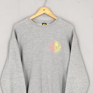 May include: A gray sweatshirt with a crew neck and long sleeves. The sweatshirt features a circular graphic on the chest with a hand design in pink, yellow, and orange. The sweatshirt is hanging on a wooden hanger.