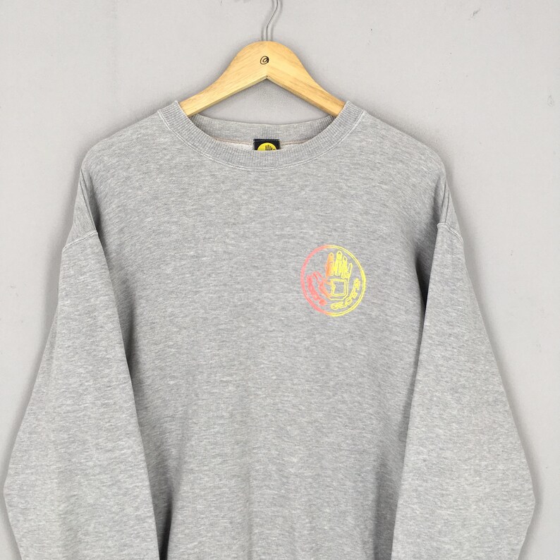 May include: A gray sweatshirt with a crew neck and long sleeves. The sweatshirt features a circular graphic on the chest with a hand design in pink, yellow, and orange. The sweatshirt is hanging on a wooden hanger.