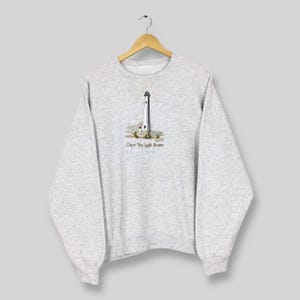May include: A light gray sweatshirt featuring a detailed illustration of a lighthouse with the text "Cape May Light House" below the image. The lighthouse is white with a black top, set against a backdrop of green and brown foliage.