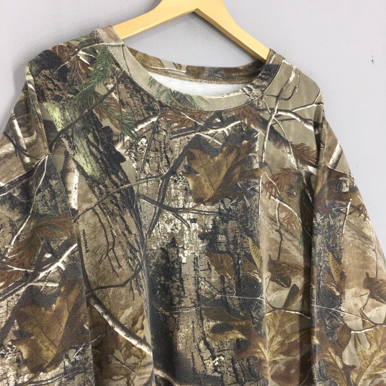 Op de afbeelding: Een sweatshirt met camouflagepatroon hangt aan een houten hanger. De top met lange mouwen heeft een mix van bruine, groene en bruine tinten, die een bosrijke omgeving nabootsen. De crew neck sweatshirt is gemaakt van een zacht materiaal.