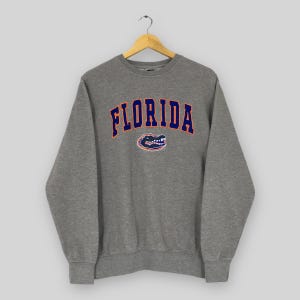 May include: A heather gray sweatshirt with the word "FLORIDA" in navy blue arched lettering with orange trim. Below the text is a navy blue and orange alligator logo. The sweatshirt has a crew neck and long sleeves.
