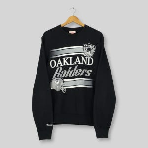 Può includere: Felpa girocollo nera con la scritta "OAKLAND Raiders" in bianco e strisce argentate e grigie. Il logo dei Raiders è in alto a destra. Abbigliamento sportivo vintage.