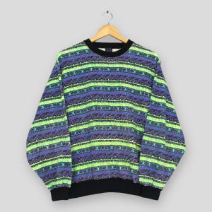 Könnte beinhalten: Ein Vintage-Sweatshirt mit Rundhalsausschnitt und einem lebendigen Muster. Das langärmlige Oberteil hat horizontale Streifen in Neongrün, Lila und Blau mit schwarzen Bündchen und Kragen. Das Sweatshirt hängt an einem Holzkleiderbügel.