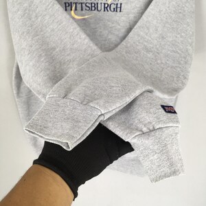 May include: A gray sweatshirt with the text "UNIVERSITY OF PITTSBURGH" in navy blue and yellow. The sweatshirt has ribbed cuffs and a small blue and white patch on the sleeve.