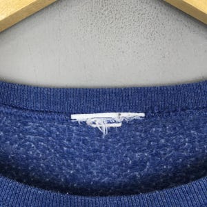 May include: A blue crewneck sweatshirt featuring a textured, heathered fabric finish. The garment includes a ribbed collar neckline and a white, frayed, and worn-out rectangular clothing label tag positioned at the center of the inner collar.