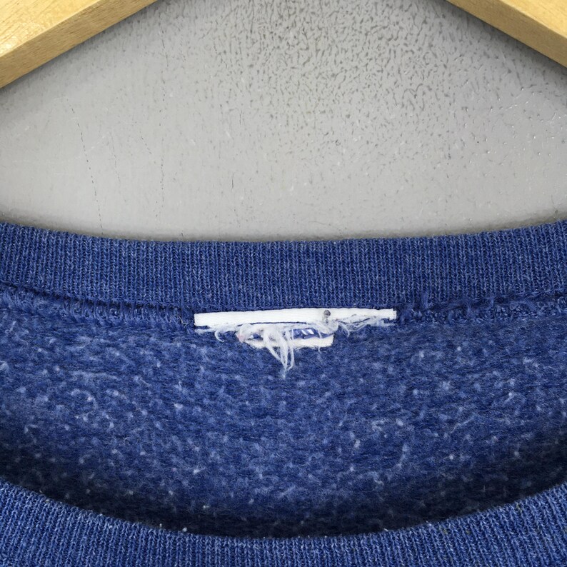 May include: A blue crewneck sweatshirt featuring a textured, heathered fabric finish. The garment includes a ribbed collar neckline and a white, frayed, and worn-out rectangular clothing label tag positioned at the center of the inner collar.