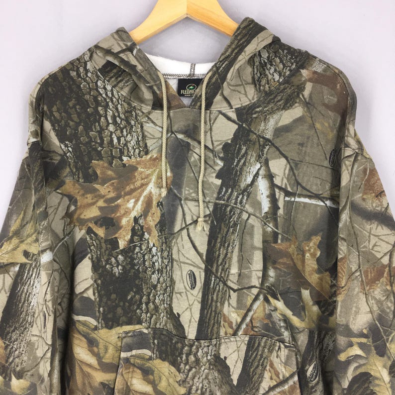 Peut inclure: Sweat &agrave; capuche camouflage avec une poche avant et un cordon de serrage. Le motif pr&eacute;sente des nuances de marron, de vert et de beige, ressemblant &agrave; une sc&egrave;ne de for&ecirc;t. Le sweat est suspendu &agrave; un cintre en bois.