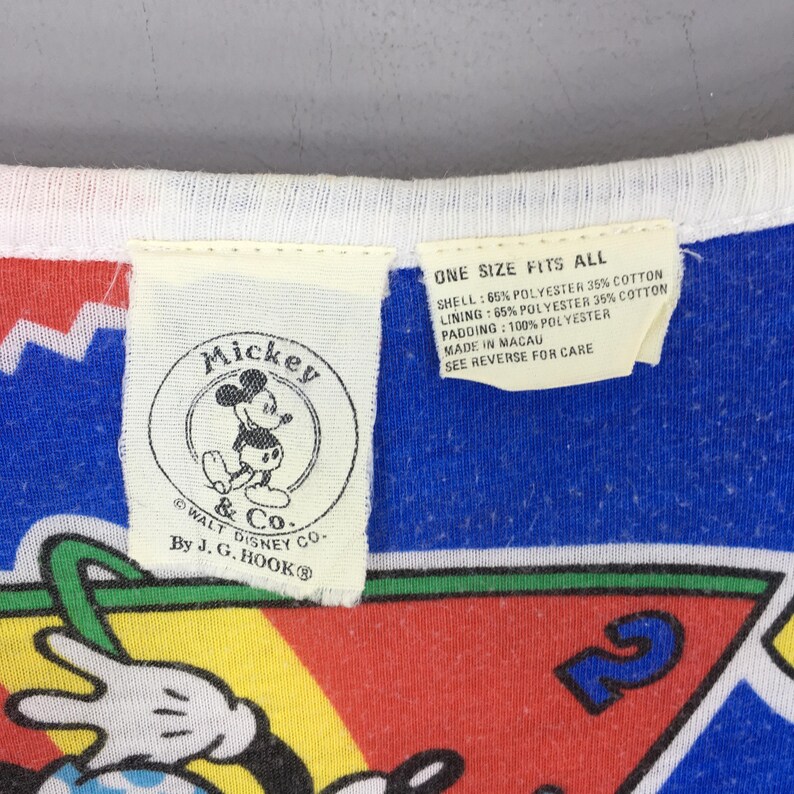 May include: A garment featuring a white tag with a circular Mickey & Co. by J.G. Hook logo and a second tag stating One Size Fits All, 65% polyester, 35% cotton, and 100% polyester padding. The fabric displays a colorful pattern with blue, red, and yellow.