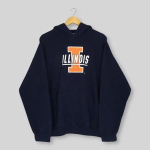 May include: A navy blue hoodie with the word "ILLINOIS" in white and orange block letters on the front. It features a front pocket and a drawstring hood. The garment is suspended from a wooden hanger.