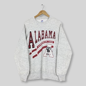 May include: A light gray crewneck sweatshirt featuring the word ALABAMA in dark red letters with black outlines. Below the text, a red ribbon graphic displays CRIMSON TIDE, positioned above a large white letter A with a black elephant illustration.