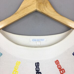May include: White crew neck sweater with colorful embroidered text. The text is in various colors, including yellow, blue, and red. The sweater is hanging on a wooden hanger.