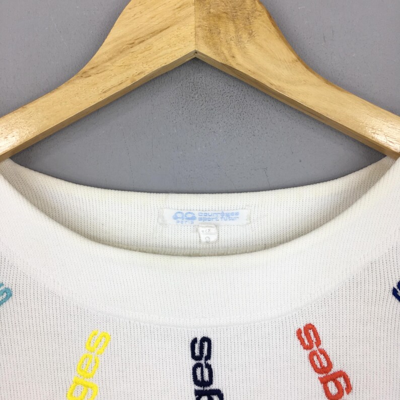 May include: White crew neck sweater with colorful embroidered text. The text is in various colors, including yellow, blue, and red. The sweater is hanging on a wooden hanger.