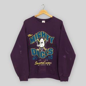 Op de afbeelding: Een vintage paarse sweatshirt met de tekst "The Mighty Ducks" in geel en teal, met een hockeymasker-afbeelding. De sweatshirt bevat ook de tekst "Inaugural Season" en "California".