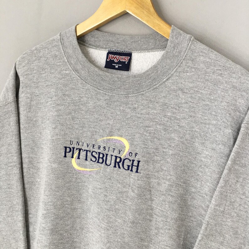 May include: A gray crew neck sweatshirt with the text "UNIVERSITY OF PITTSBURGH" in navy blue and yellow. The sweatshirt has a small "Jansport" tag at the neckline. The sweatshirt is hanging on a wooden hanger.