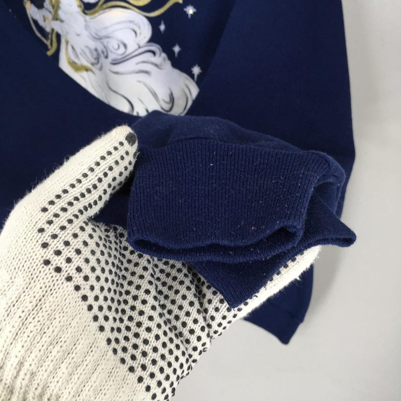 May include: A dark navy blue long-sleeve sweatshirt featuring a graphic print of a white-haired figure with gold accents and small white star details. The garment has a ribbed knit cuff at the end of the sleeve and a crew neckline design.
