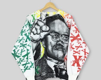 May include: White long-sleeve sweatshirt featuring a black and white image of a man wearing glasses, pointing. The sleeves have green, red, and orange accents with the letter "X" and the name "Malcolm" printed on them. The sweatshirt is hanging on a wooden hanger.