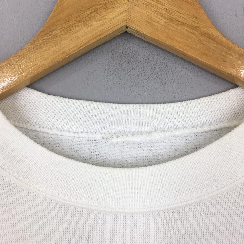 May include: Close-up of a white crew neck sweatshirt collar. The collar is ribbed and has a slightly distressed appearance. The sweatshirt is hanging on a wooden hanger against a gray background.