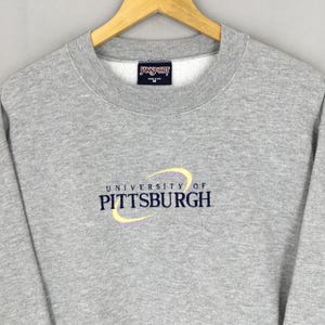 May include: A gray crewneck sweatshirt with the "University of Pittsburgh" logo embroidered in yellow and navy blue. The sweatshirt has a "Jansport" tag at the neckline. The garment is made of a soft, comfortable material.