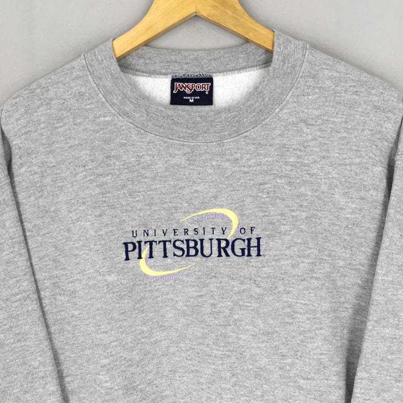 May include: A gray crewneck sweatshirt with the "University of Pittsburgh" logo embroidered in yellow and navy blue. The sweatshirt has a "Jansport" tag at the neckline. The garment is made of a soft, comfortable material.