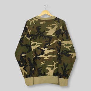 May include: A camouflage-patterned sweatshirt in shades of green, brown, and tan. The long-sleeved top has a crew neck and ribbed cuffs and hem. The garment is hanging on a wooden hanger against a light gray background.