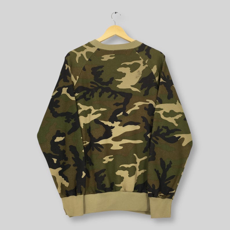 May include: A camouflage-patterned sweatshirt in shades of green, brown, and tan. The long-sleeved top has a crew neck and ribbed cuffs and hem. The garment is hanging on a wooden hanger against a light gray background.