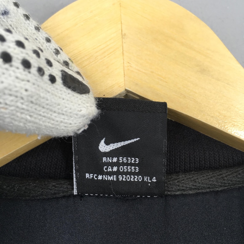 May include: A white ceramic mug with a black handle and the text "But First, Coffee" in a bold, black font. The mug is set against a blurred background. The mug appears to be approximately 10 cm tall.