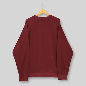 Może przedstawiać: Bordowa bluza z długimi rękawami, wisząca na drewnianym wieszaku. Bluza z okrągłym dekoltem jest w jednolitym kolorze i wydaje się być wykonana z miękkiego materiału. Odzież jest prezentowana na jasnoszarym tle.